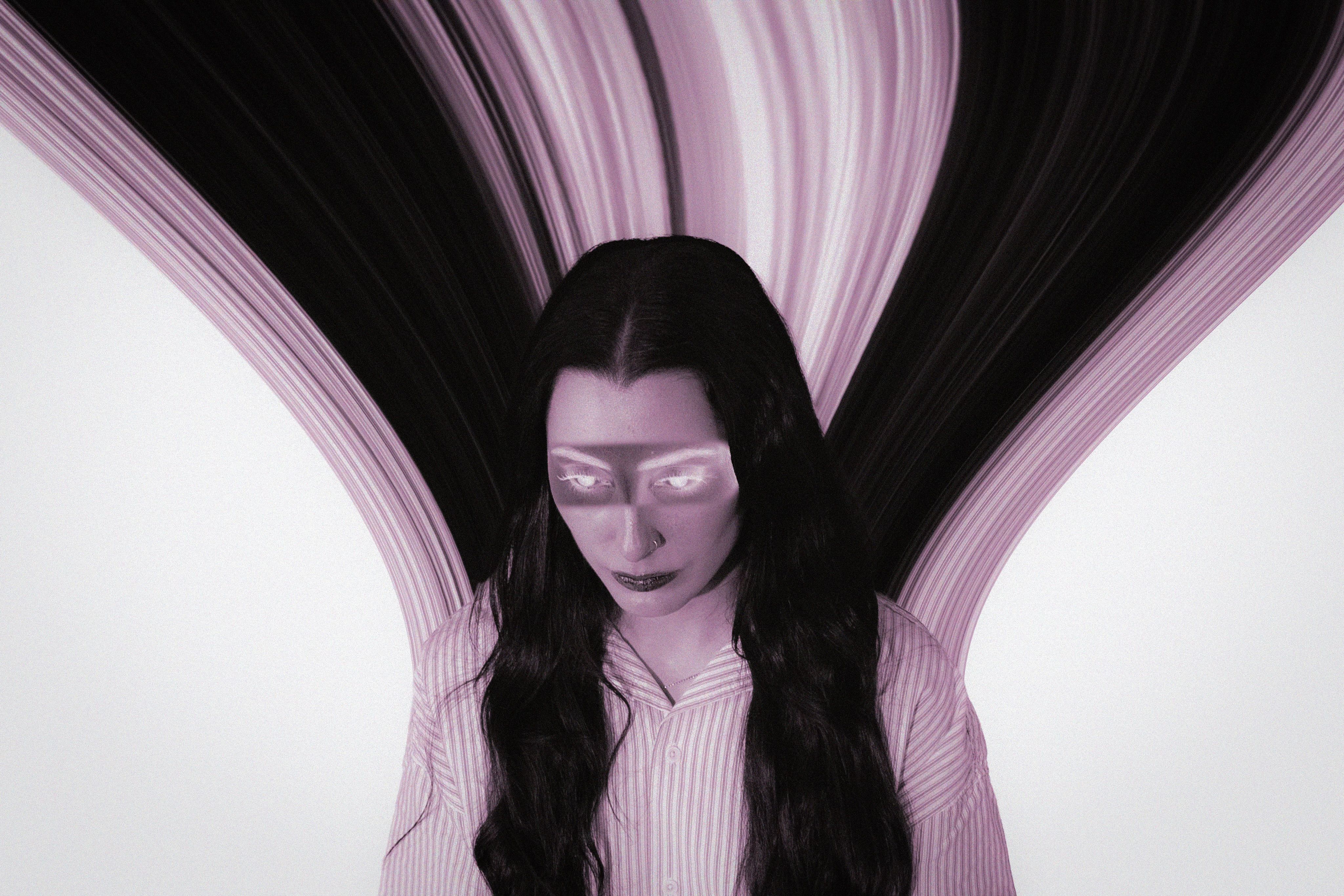 A high-contrast portrait in green and pink light, a woman looking downward with dramatic dark makeup and a soft glow around her.