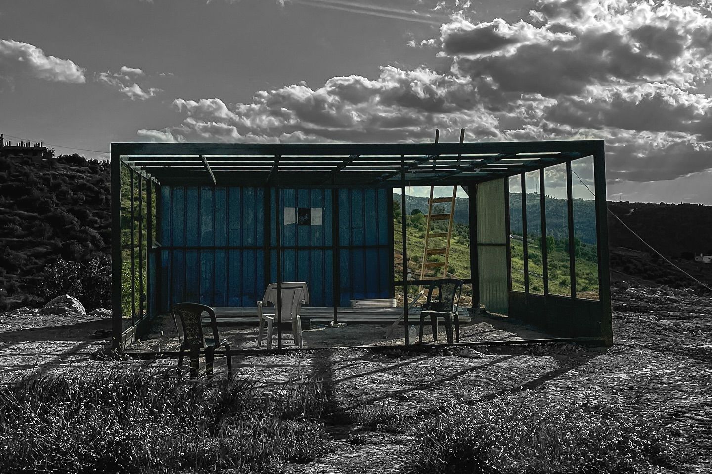 A black-and-white photo of an unfinished glass-and-metal structure with two white plastic chairs inside it, standing alone on a hillside under a dramatic cloudy sky.