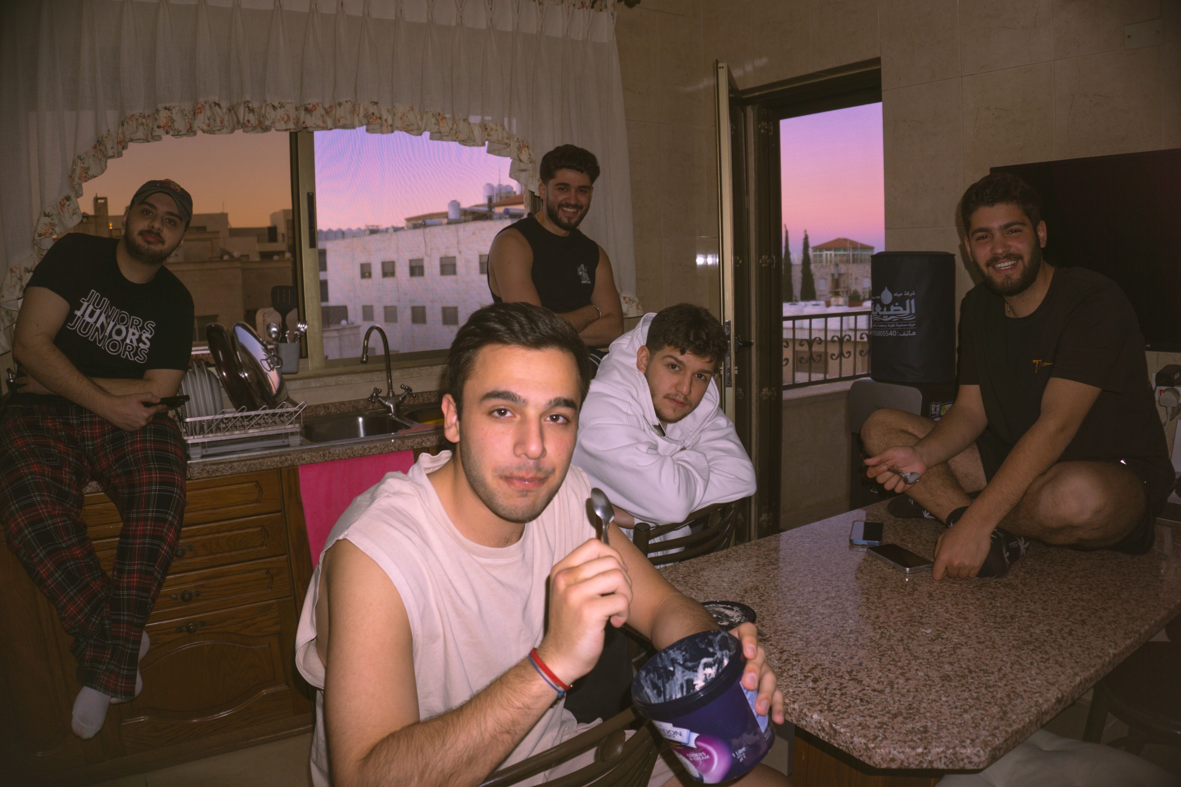 Five young men gathered in a kitchen at sunset, one in the foreground holding a spoon and a tub of ice cream.