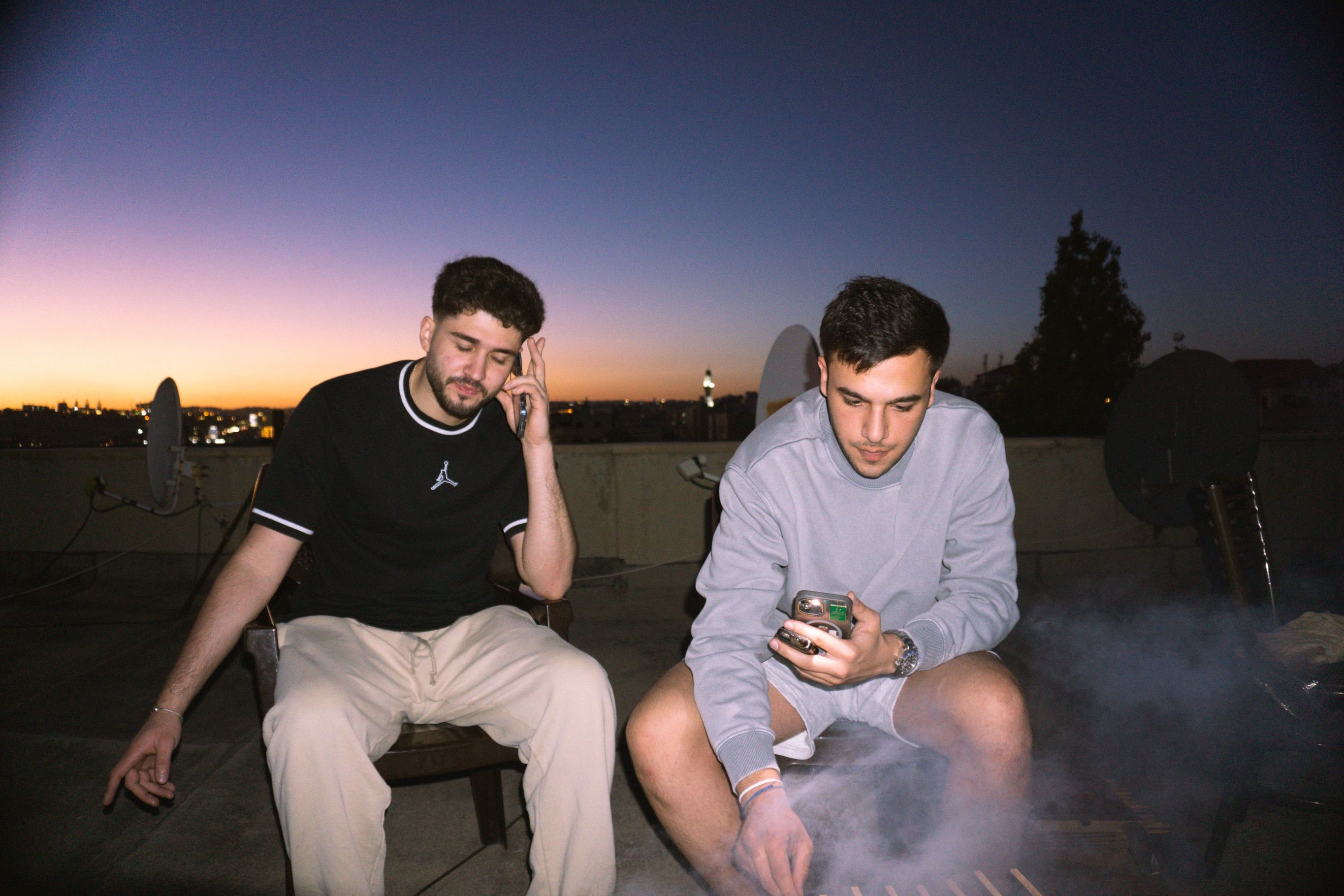 Two young men sit on a rooftop at dusk, satellite dishes and a city skyline behind them, one holding a phone to his ear.