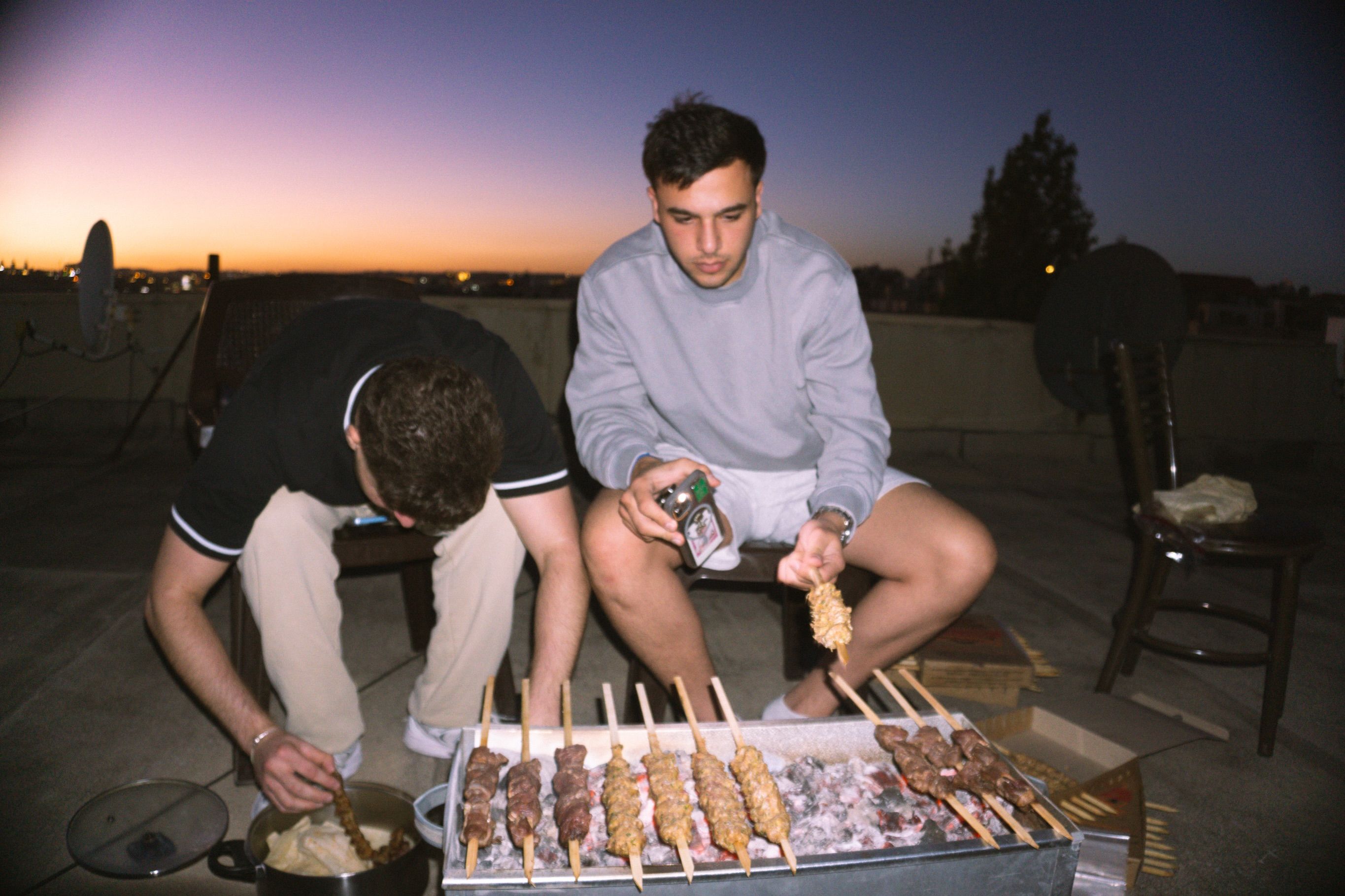 Two young men grill skewered meat over hot coals on a rooftop at dusk, a barbecue tray between them.