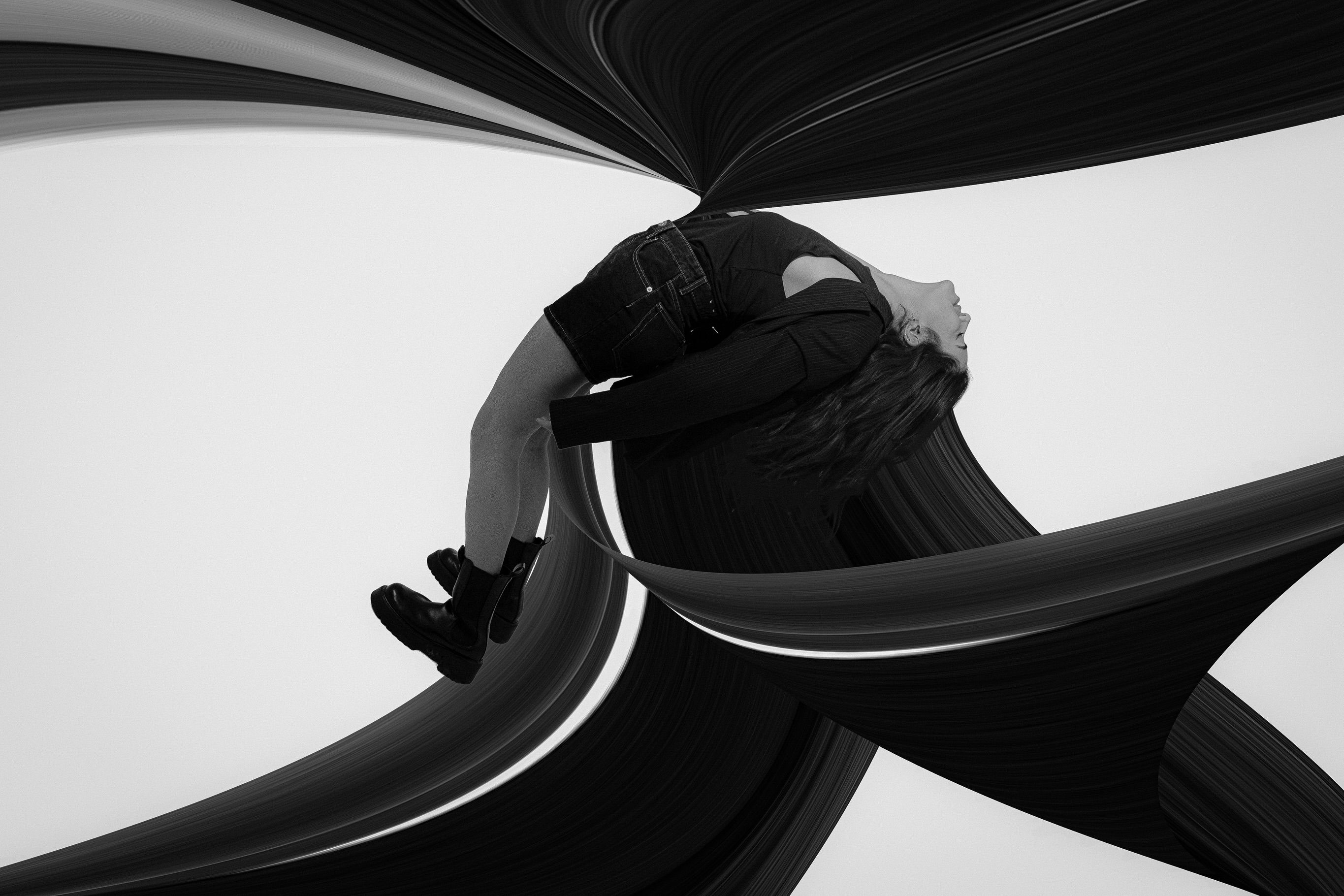 A black-and-white long-exposure photo of a person bent backward, surrounded by swirling dark ribbon-like trails of motion.