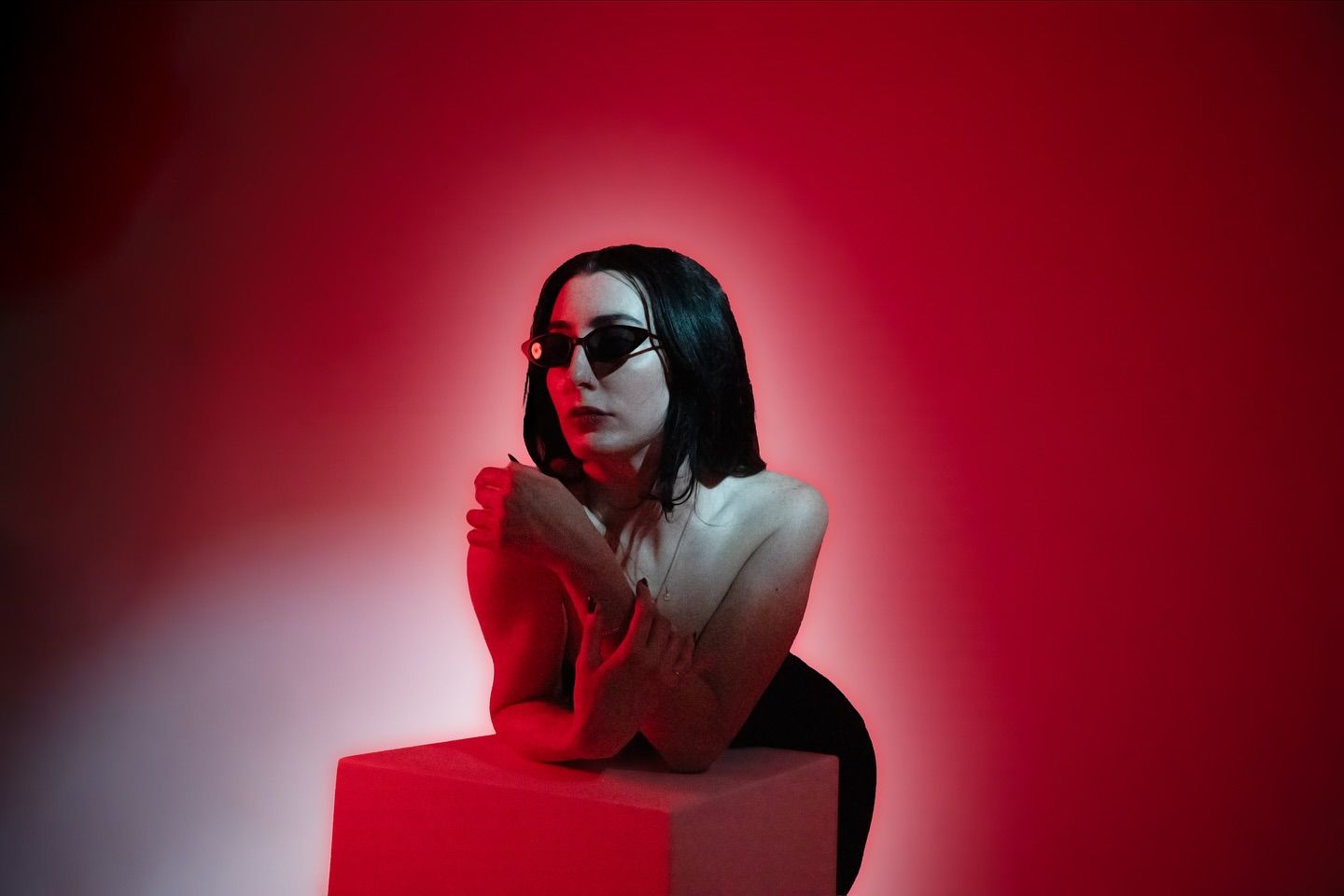 A woman in dark sunglasses leans on a white cube, arms crossed, lit in deep red studio light.
