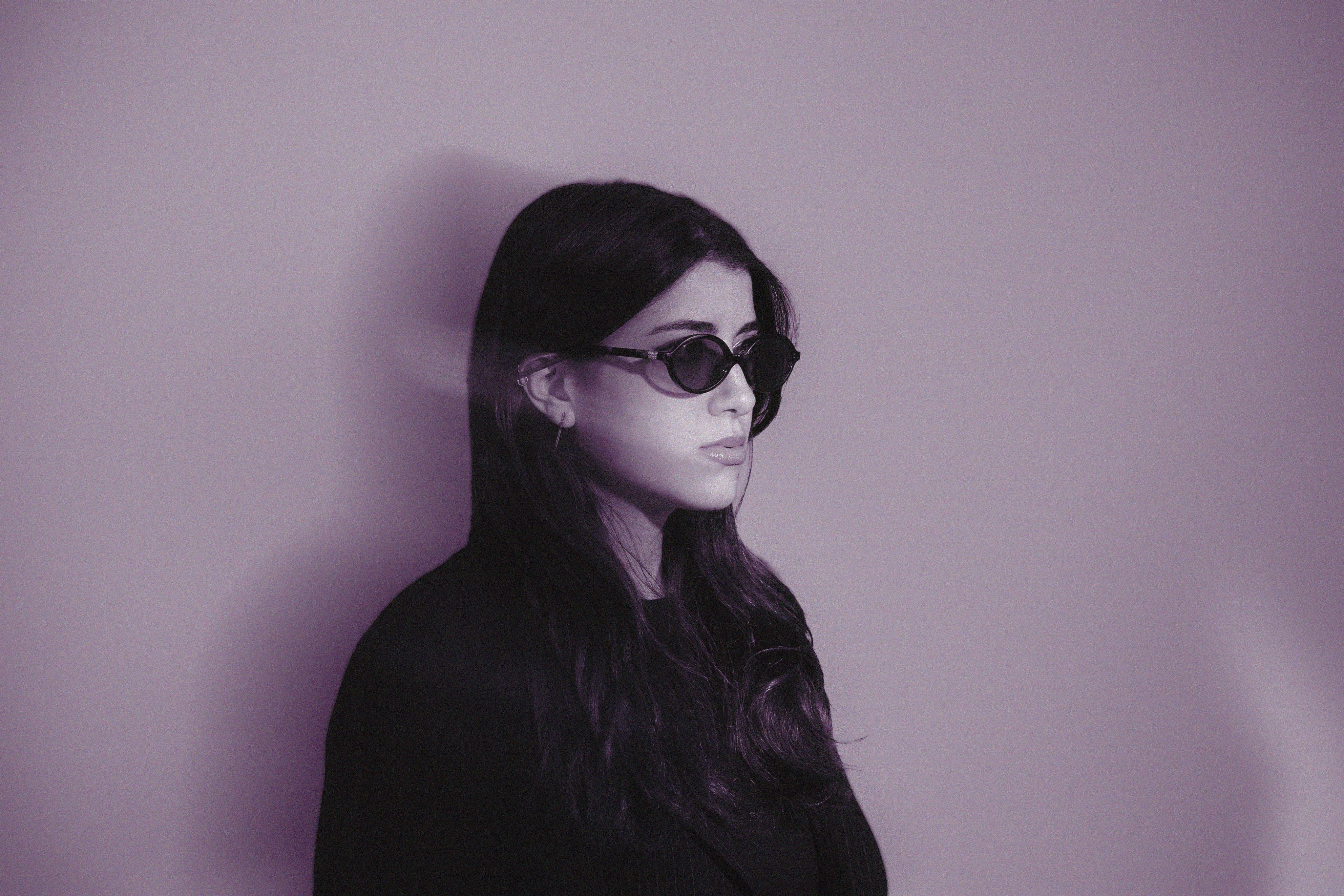 A profile portrait of a woman in dark cat-eye sunglasses, lit in violet tones against a plain wall.